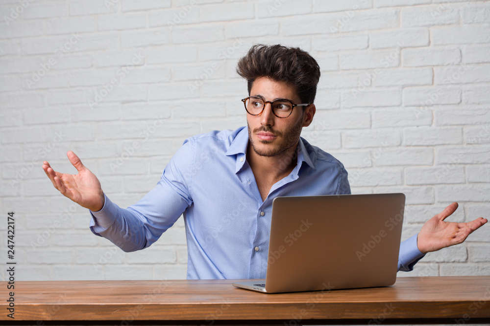 Young business man sitting and working on a laptop doubting and shrugging shoulders, concept of indecision and insecurity, uncertain about something Young business man sitting and working on a laptop doubting and shrugging shoulders, concept of indecision and insecurity, uncertain about something