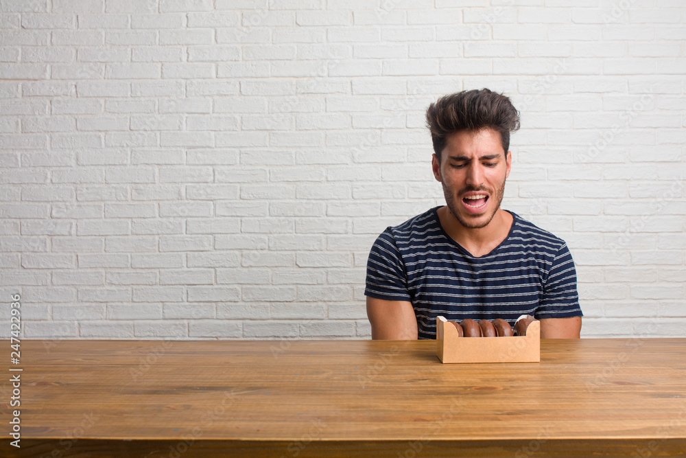 Young handsome and natural man sitting on a table screaming angry, expression of madness and mental instability, open mouth and half-opened eyes, madness concept. Eating chocolate doughnuts. Young handsome and natural man sitting on a table screaming angry, expression of madness and mental instability, open mouth and half-opened eyes, madness concept. Eating chocolate doughnuts.