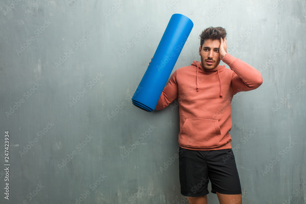 Young fitness man against a grunge wall frustrated and desperate, angry and sad with hands on head. Holding a blue mat for practicing yoga.