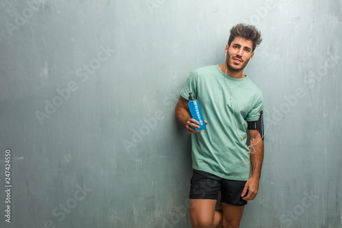 Young fitness man against a grunge wall cheerful and with a big smile, confident, friendly and sincere, expressing positivity and success. Holding a blue energy drink.