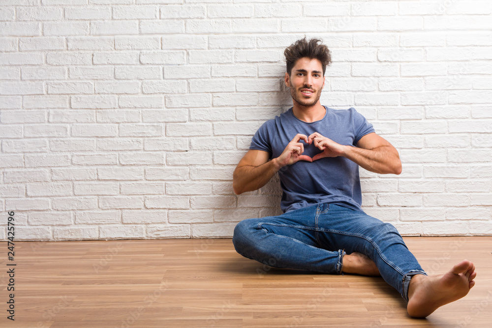 Young natural man sit on a wooden floor making a heart with hands, expressing the concept of love and friendship, happy and smiling