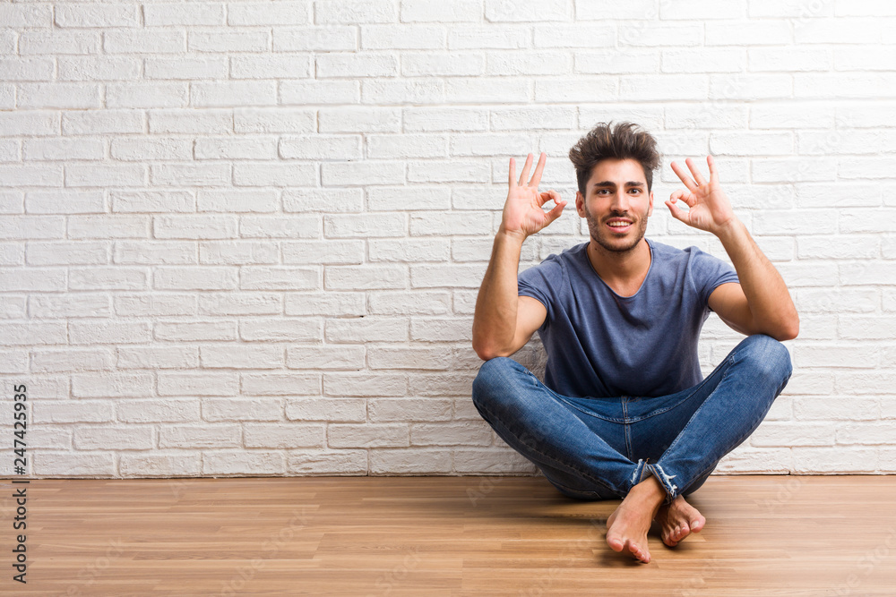 Young natural man sit on a wooden floor cheerful and confident doing ok gesture, excited and screaming, concept of approval and success Young natural man sit on a wooden floor cheerful and confident doing ok gesture, excited and screaming, concept of approval and success
