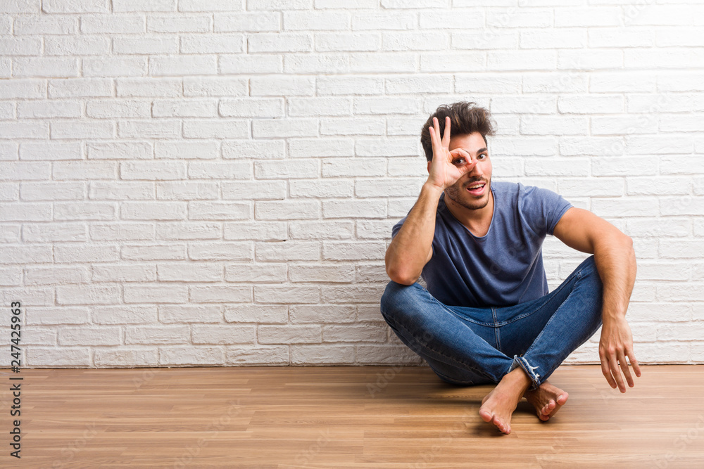 Young natural man sit on a wooden floor cheerful and confident doing ok gesture, excited and screaming, concept of approval and success
