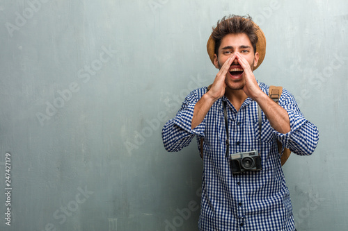 Young handsome traveler man wearing a straw hat, a backpack and a photo camera screaming happy, surprised by an offer or a promotion, gaping, jumping and proud