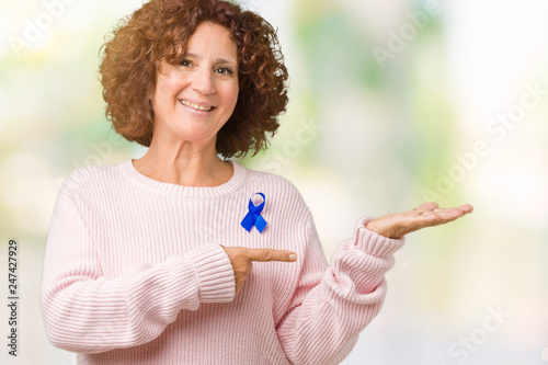 Middle ager senior woman wearing changeable blue color ribbon awareness over isolated background amazed and smiling to the camera while presenting with hand and pointing with finger.