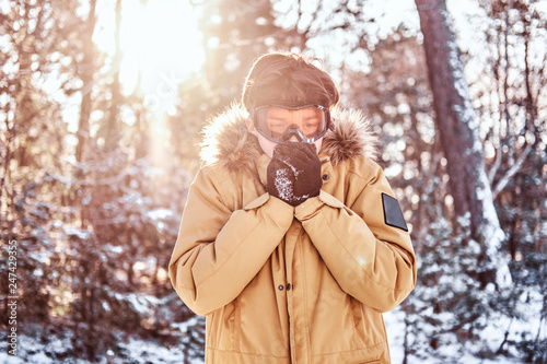 Young snowboarder dressed in warm clothes and goggles, warms his hands standing in the snowy forest during sunrise