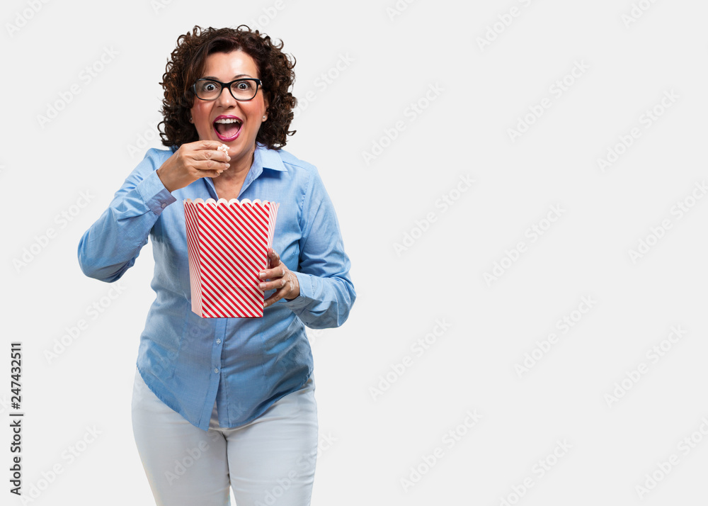 Middle aged woman happy and fascinated, holding a striped popcorn bucket, surprised by the new movie, eyes open and expression of admiration Middle aged woman happy and fascinated, holding a striped popcorn bucket, surprised by the new movie, eyes open and expression of admiration