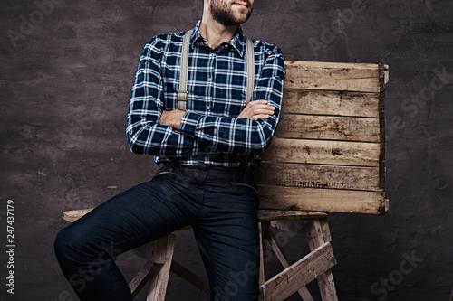 Old-fashioned man wearing a checkered shirt with suspenders with his arms crossed sitting on a wooden scaffolding