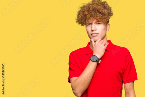 Young handsome man with afro hair wearing red t-shirt looking confident at the camera with smile with crossed arms and hand raised on chin. Thinking positive.