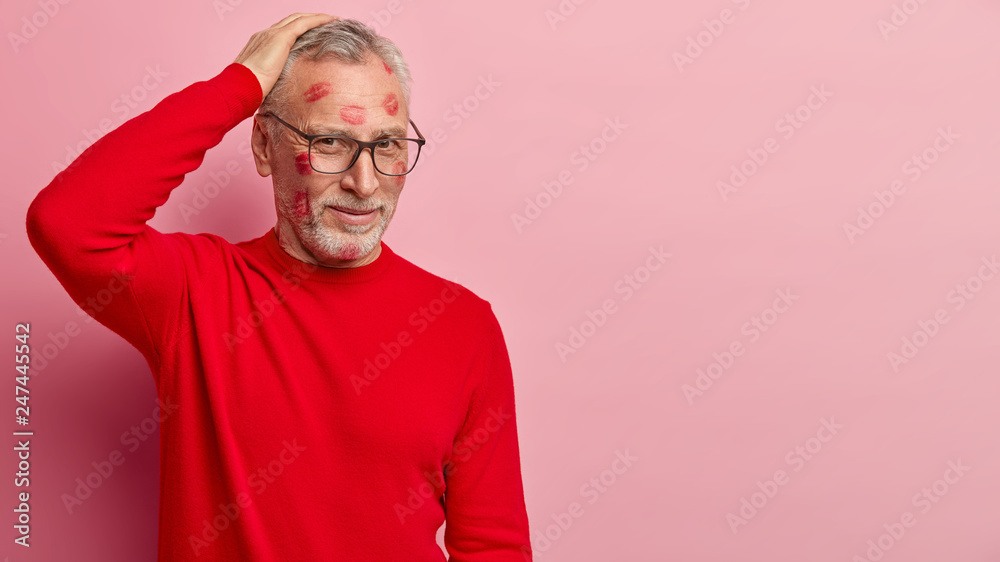 Attractive unshaven male pensioner full of lipstick marks of kisses, wears optical glasses and red jumper, poses over pink studio wall with empty space for advertising content. Valentines Day concept Attractive unshaven male pensioner full of lipstick marks of kisses, wears optical glasses and red jumper, poses over pink studio wall with empty space for advertising content. Valentines Day concept
