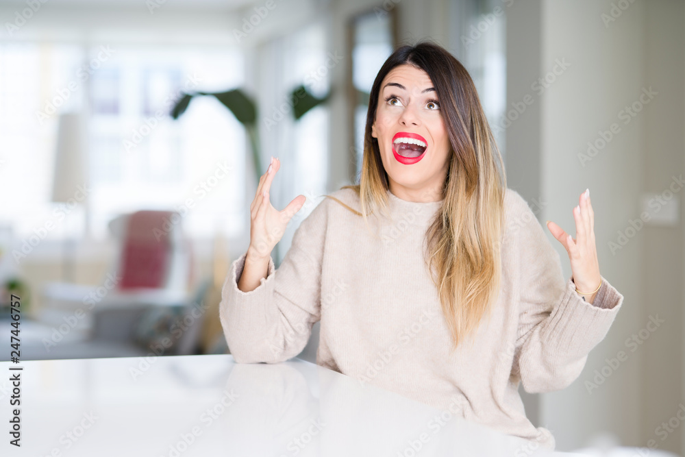 Young beautiful woman wearing winter sweater at home crazy and mad shouting and yelling with aggressive expression and arms raised. Frustration concept. Young beautiful woman wearing winter sweater at home crazy and mad shouting and yelling with aggressive expression and arms raised. Frustration concept.