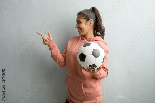 Young sporty indian woman against a wall pointing to the side, smiling surprised presenting something, natural and casual. Holding a soccer ball to play a game. © Asier