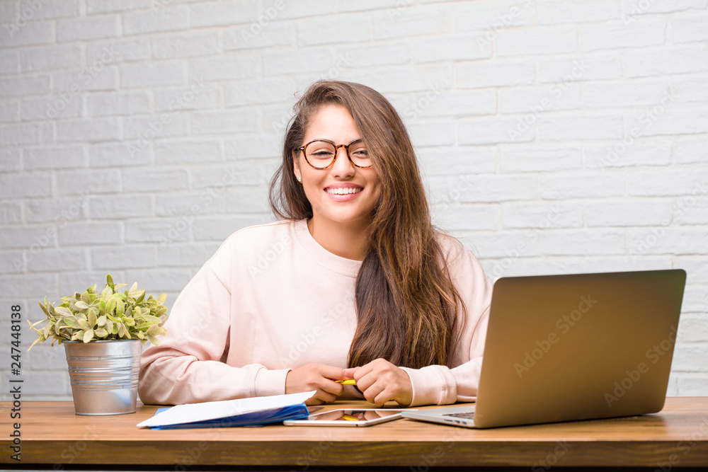 Portrait of young student latin woman sitting on her desk cheerful and with a big smile, confident, friendly and sincere, expressing positivity and success Portrait of young student latin woman sitting on her desk cheerful and with a big smile, confident, friendly and sincere, expressing positivity and success