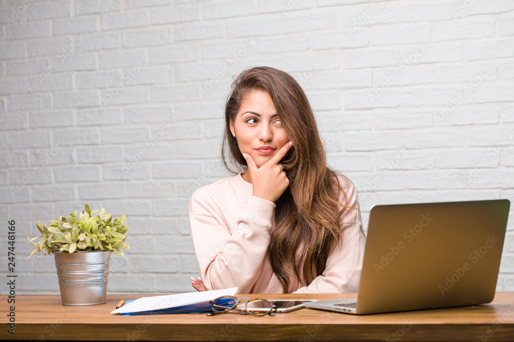 Portrait of young student latin woman sitting on her desk thinking and looking up, confused about an idea, would be trying to find a solution Portrait of young student latin woman sitting on her desk thinking and looking up, confused about an idea, would be trying to find a solution