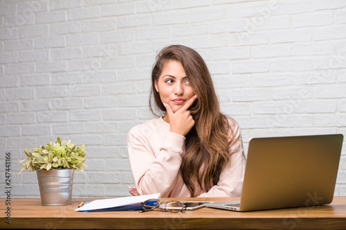 Portrait of young student latin woman sitting on her desk thinking and looking up, confused about an idea, would be trying to find a solution