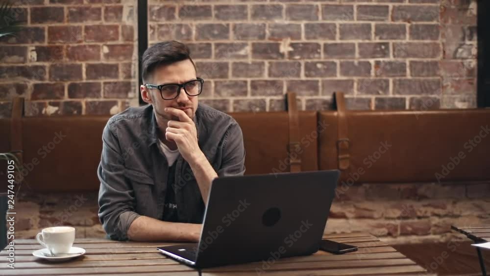 Pensive brunette man in eyewear wearing gray shirt looking away and thinking about something while sitting at the table with laptop in comfortable cafe