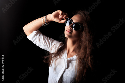 The cool girl in black sunglasses. Portrait of pretty young woman with long curly wavy brown hair on the black background.