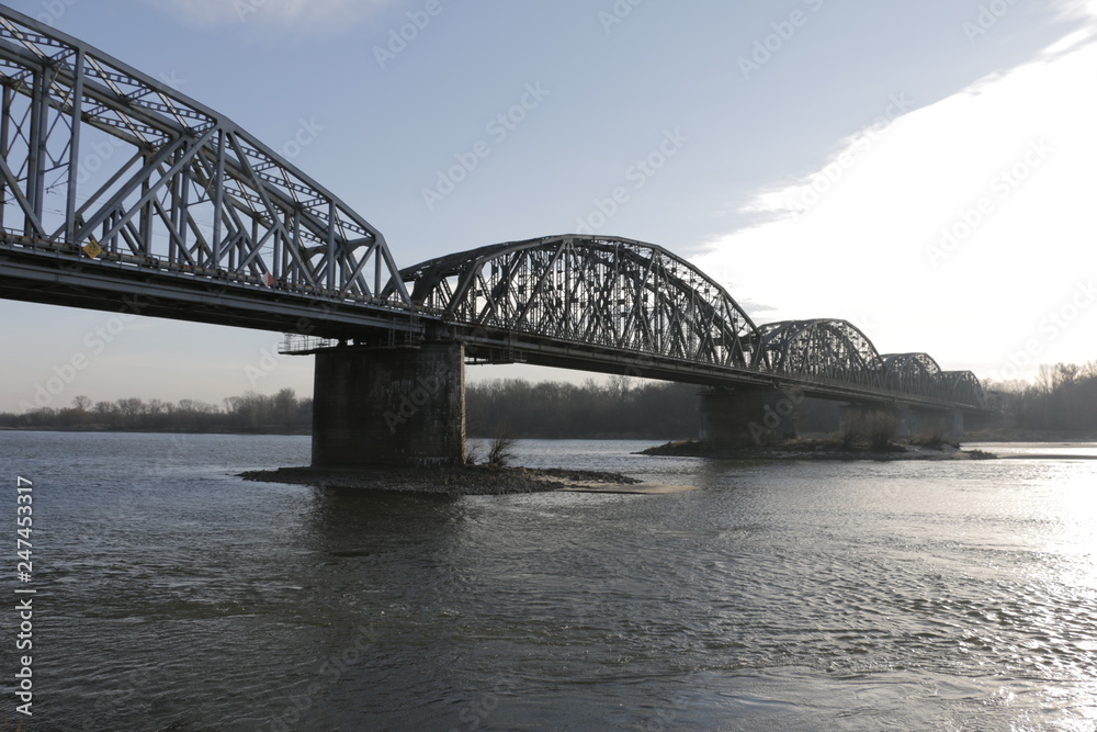 The Ernest Malinowski Bridge in Torun. Railway bridge on the Vistula river. Visible spans, steel trusses, supports, water current. A winter, cold, windy day. A shore with trees in the background.