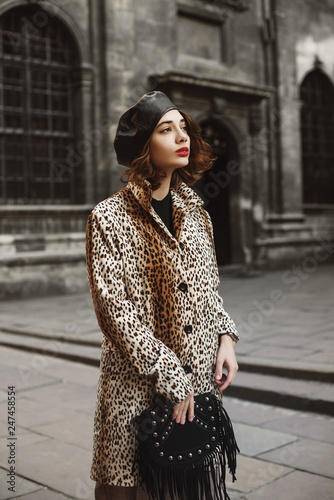 Outdoor fashion portrait of young woman wearing trendy leopard print coat, leather beret, holding black suede bag with fringe, posing in street of European city