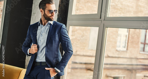 Confident and stylish. Businessman in sunglasses and fashion suit is thoughtfully looking in window while standing at office
