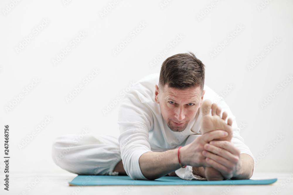 Handsome young man in white clothes does exercises. Yoga asanas and poses for stretching and meditating on a blue rug. Against the background of a white wall. Handsome young man in white clothes does exercises. Yoga asanas and poses for stretching and meditating on a blue rug. Against the background of a white wall.