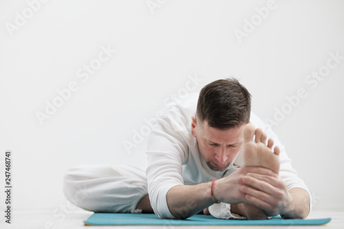 Handsome young man in white clothes does exercises. Yoga asanas and poses for stretching and meditating on a blue rug. Against the background of a white wall.