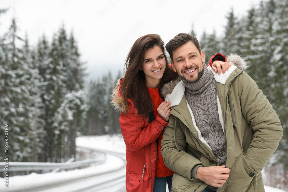 Couple in warm clothes near snowy forest, space for text. Winter vacation