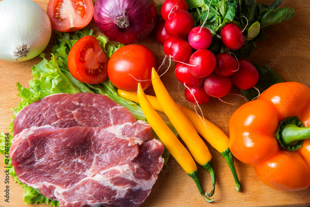 Pork-neck meat steaks on lettuce on background of radishes, tomato, red chili peppers, yellow chili peppers, green paprika, yellow paprika, red paprika, black pepper. Horizontal. Red onions, shallots Pork-neck meat steaks on lettuce on background of radishes, tomato, red chili peppers, yellow chili peppers, green paprika, yellow paprika, red paprika, black pepper. Horizontal. Red onions, shallots