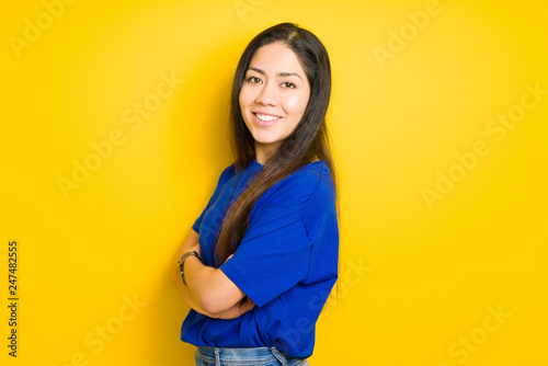 Beautiful young brunette woman standing with crossed arms and smiling over yellow isolated background