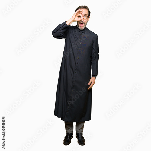 Middle age priest man wearing catholic robe doing ok gesture shocked with surprised face, eye looking through fingers. Unbelieving expression.