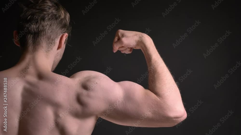 Close-up rear portrait of young caucasian man showing his muscular biceps and demonstrating how strong it is on black background. Close-up rear portrait of young caucasian man showing his muscular biceps and demonstrating how strong it is on black background.