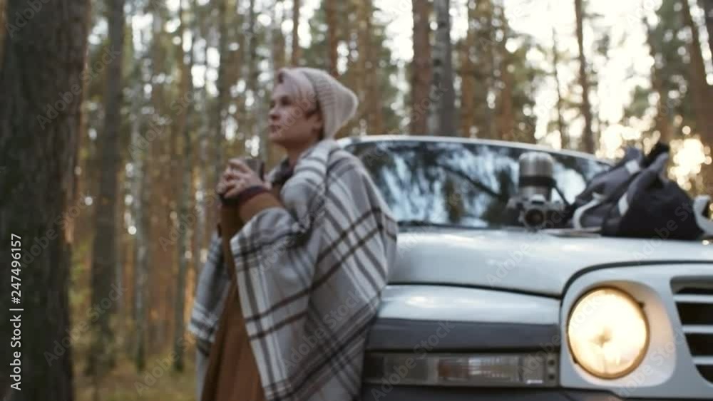 Zoom in shot of relaxed young Caucasian woman in beanie standing by crossover car and enjoying warm drink in forest Zoom in shot of relaxed young Caucasian woman in beanie standing by crossover car and enjoying warm drink in forest
