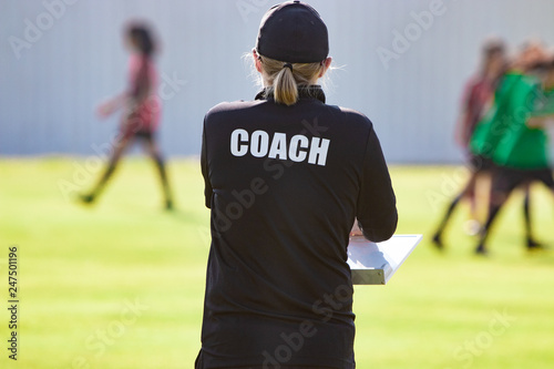 Back view of female football coach in black COACH shirt at an outdoor sport field watching her team play