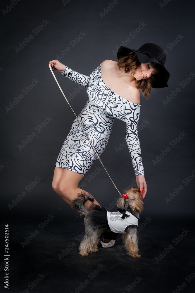 young beautiful woman in black hat with yorkshire terrier on leash in photo studio stands on black background