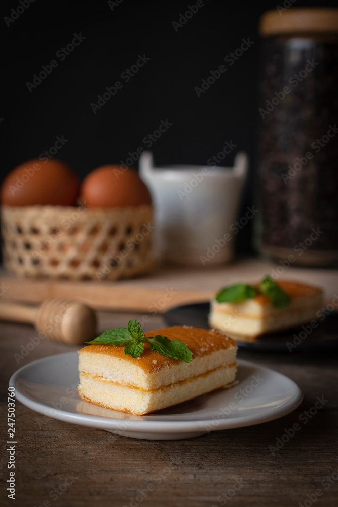 layer vanilla flavoured cake place on white plate on the wooden table there are black plate and coffee bean in glass bottles and bucket of water ceramic and basket of eggs place backside.