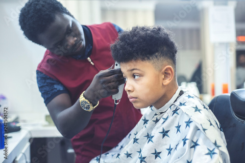 Cute african american boy in the african barbershop. Cute mixed boy makes a haircut in the African salon. Hair style. Haircut by machine for children. School boy hair style. Haircut by scissors. 