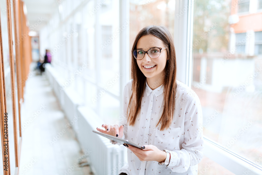 © Dusan Petkovic - Smiling Caucasian collage girl using tablet while standing in front of noticeboard and looking at camera. University hall interior.
