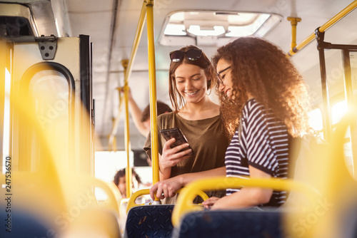 Obraz na płótnie Two girls watching phone and smiling while standing on a bus.