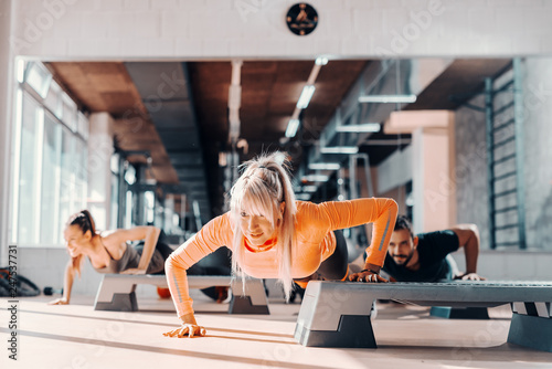 Fototapeta Naklejka Na Ścianę i Meble -  Group of sporty people doing push ups on steppers in gym. Selective focus on blonde woman, in background mirror.