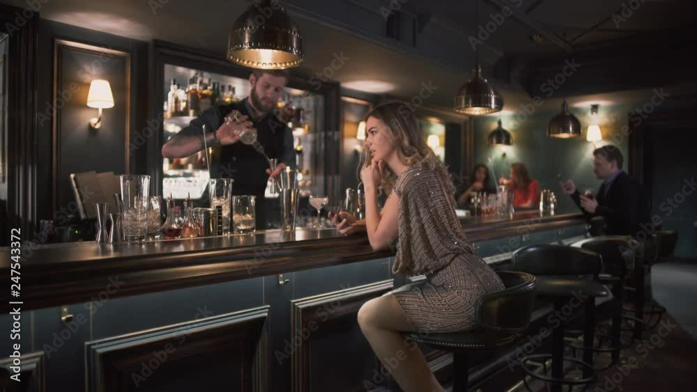 Hipster bartender combining ingredients and making cocktails in bar while young pretty woman sits near bar counter and exting message Hipster bartender combining ingredients and making cocktails in bar while young pretty woman sits near bar counter and exting message