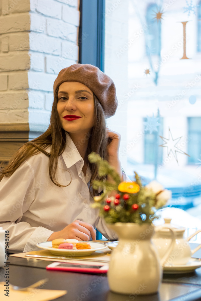 Beautiful girl in a beret sits at a table in a cafe with a cup and cake and smiles