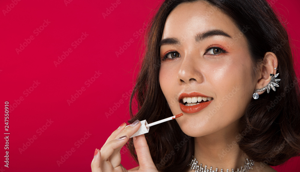 Fashion portrait of Asian Black hair tanned skin woman with strong super color red lips wearing white fur diamond earrings necklace, studio lighting red reddish background, holding liquid lipstick Fashion portrait of Asian Black hair tanned skin woman with strong super color red lips wearing white fur diamond earrings necklace, studio lighting red reddish background, holding liquid lipstick