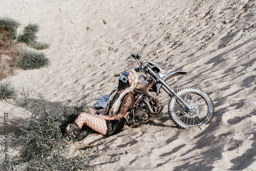 Young sexy girl biker wearing fishnet tights and mask, sitting near her silver motorcycle at sand dunes photo