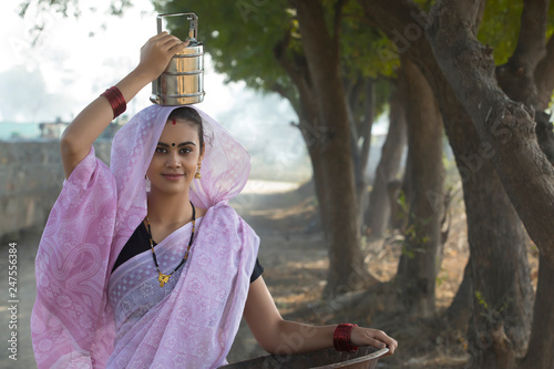 Smiling rural woman or daily wage labourer in saree carrying an iron gold pan in hand and a tiffin box on her head to work.	