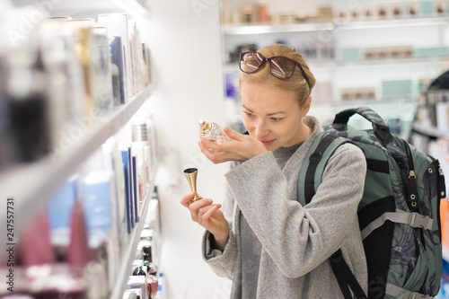 Blond young female traveler wearing coat and travel backpack choosing perfume in airport duty free store. Casual lady testing and buying cosmetics on the go in a beauty store. photo