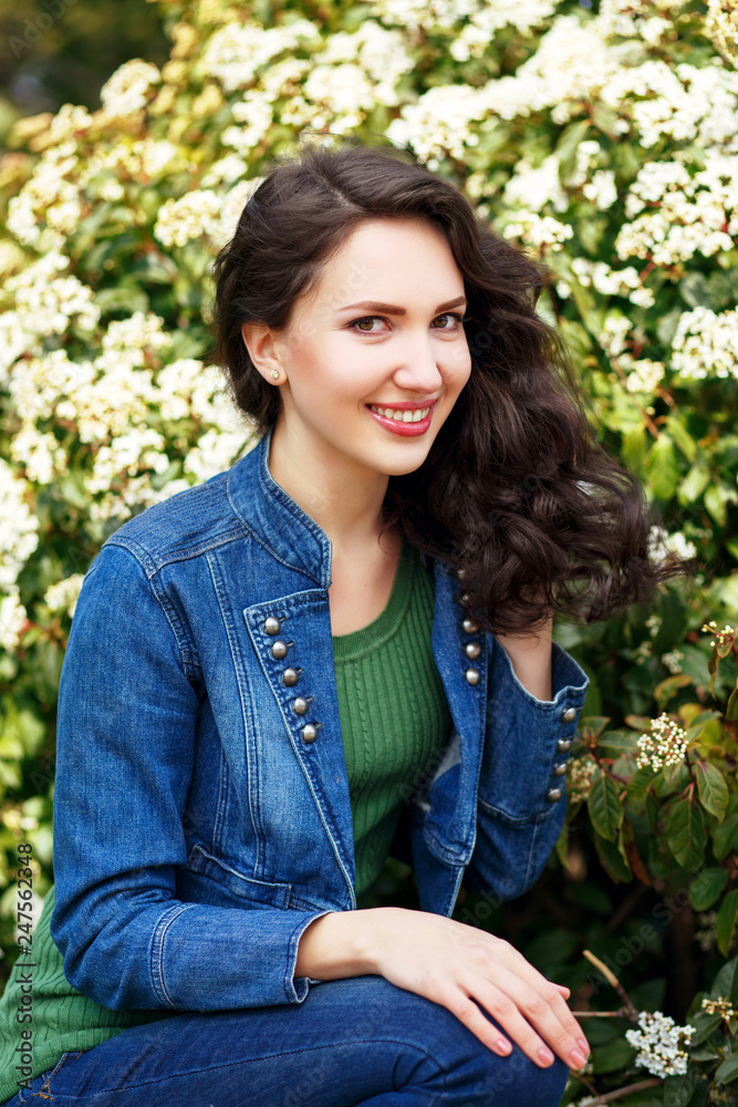 Beautiful young brunette woman sitting on the grass near white flowering blooming bushes in spring park Beautiful young brunette woman sitting on the grass near white flowering blooming bushes in spring park