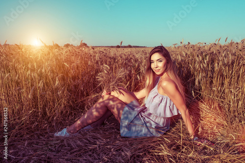 A girl in a wheat field in the summer holds wheat ears in her hand. Sits on the ground. Free space for text. Tanned skin of woman resting in the fresh air.