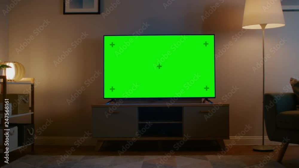 Zoom Out Shot of a TV with Horizontal Green Screen Mock Up. Cozy Evening Living Room with a Chair and Lamps Turned On at Home. Zoom Out Shot of a TV with Horizontal Green Screen Mock Up. Cozy Evening Living Room with a Chair and Lamps Turned On at Home.
