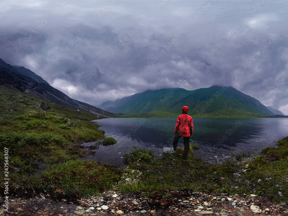 man stands by lake in red jacket in cloudy weather panorama man stands by lake in red jacket in cloudy weather panorama