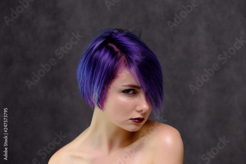 A beautiful, sexy girl with purple hair and a short haircut sits in the middle of the photo with a gray background